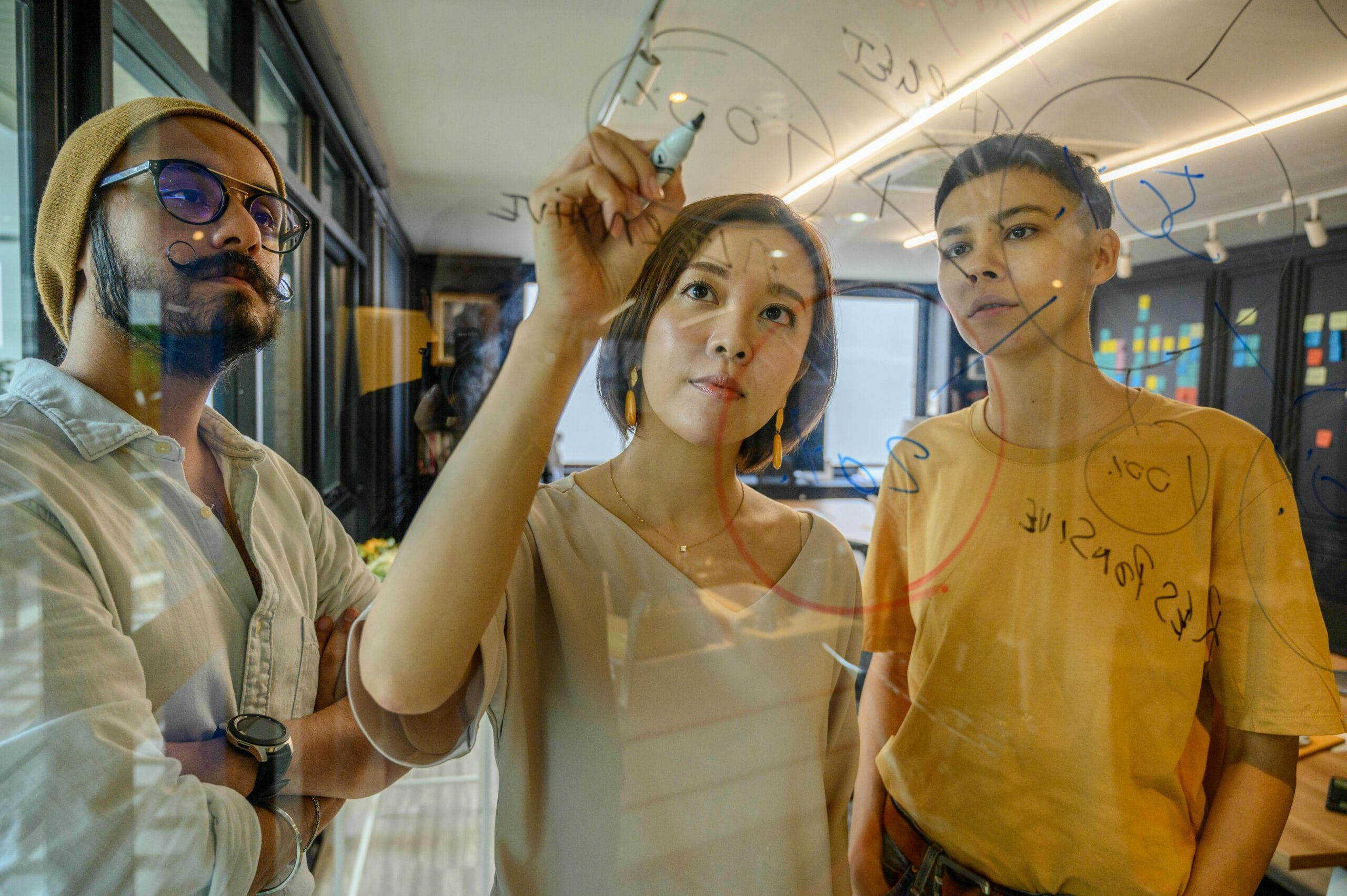 Three colleagues engaged in a strategy meeting, writing ideas on glass in a modern office.